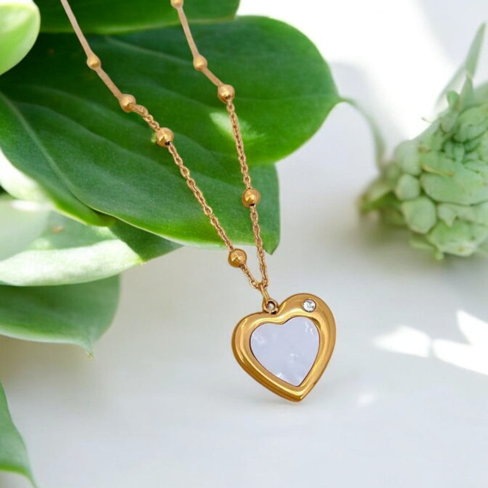 Gold heart-shaped necklace with a white heart pendant on a green leaf background